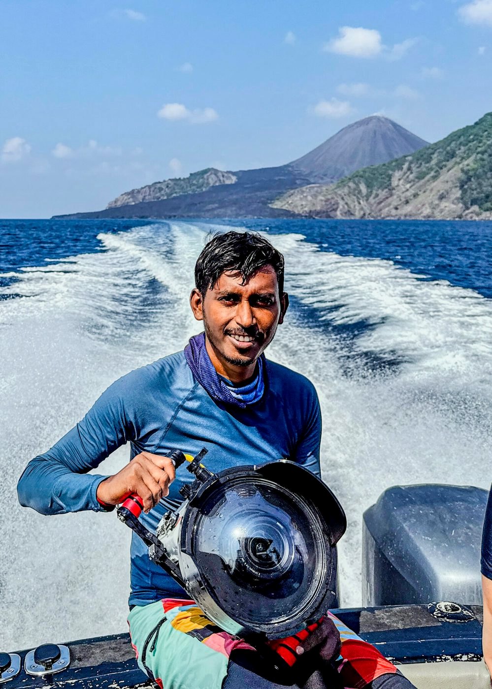 Amit Adhikari on a boat holding an underwater camera housing, with a volcano behind
